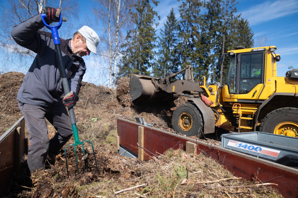 Lippalakki päässä oleva mies purkaa peräkärryn puutarhajätteitä talikolla. Taustalla näkyy keltaien kone ehostamassa kasaa.
