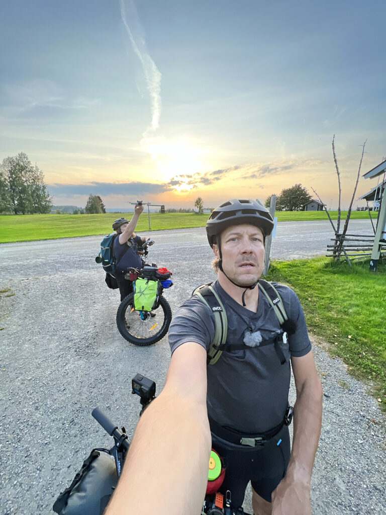 Mies ottaa selfietä pyöräilykypärä päässä. Takana näkyy pyörän vieressä toinen mies, joka laittaa ilmeisesti kameraa ilmaan.