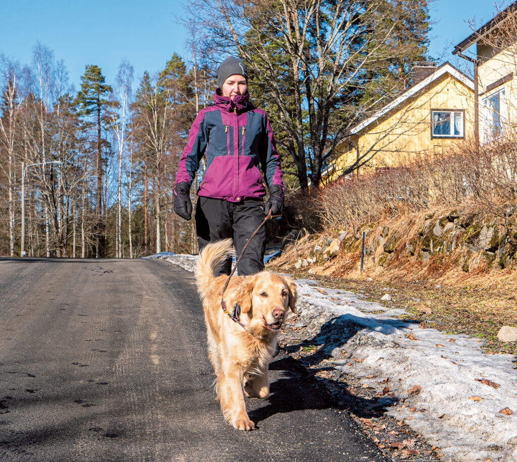 Nainen ulkoiluttaa koiraa. On kevät, koska maassa on lunta. Taustalla näkyy pari kellertävää vanhempaa omakotitaloa.