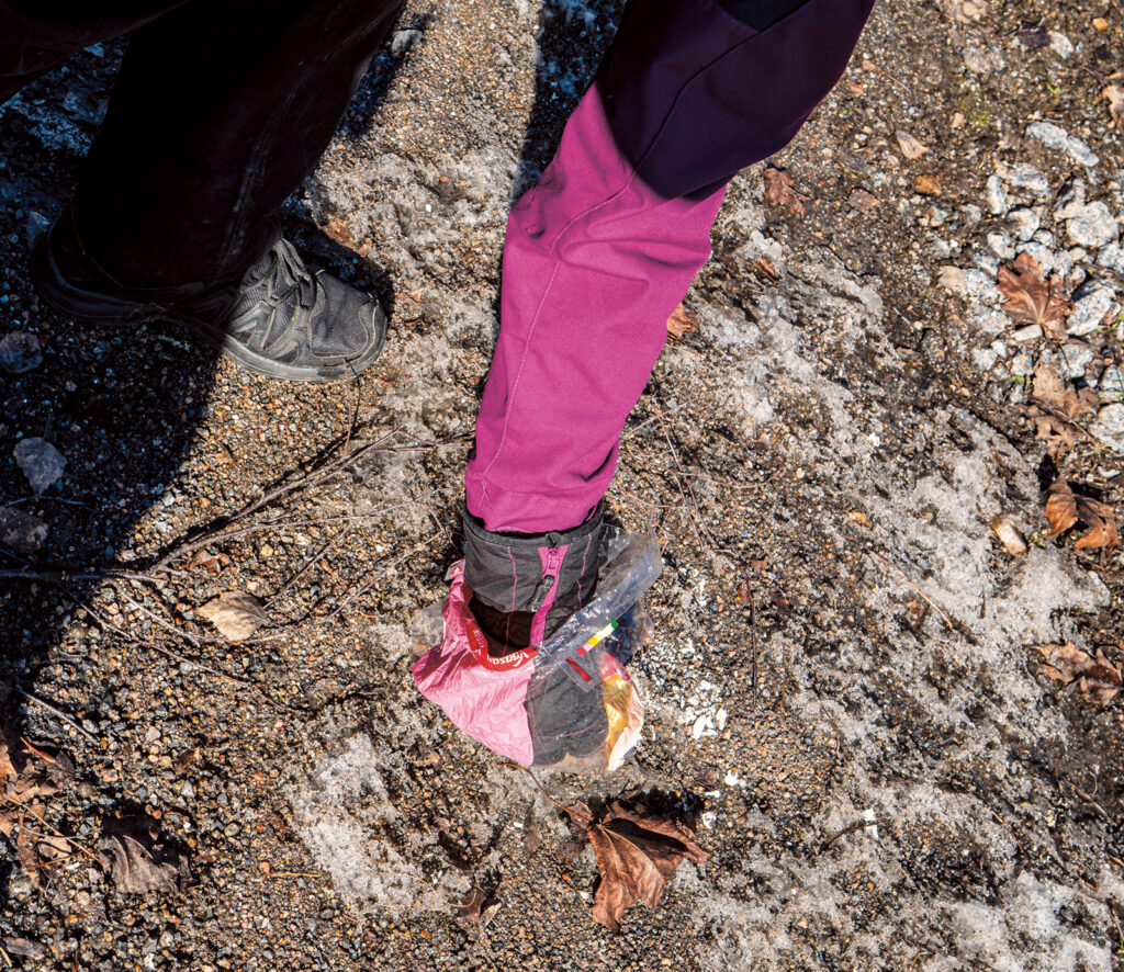 Kuvassa käsi ottaa maasta jotain, ehkä kakkaa? On kevät, sillä maassa on hieman lunta.