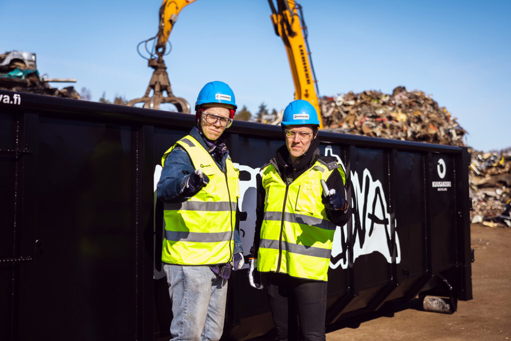 Edessä on kaksi sinikypäräistä miestä ja takana musta lava sekä metalliromua.