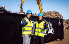 Edessä on kaksi sinikypäräistä miestä ja takana musta lava sekä metalliromua.