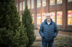 Tummansininen toppatakkinen mies seisoo ulkona. Taustalla näkyy punatiilinen rakennus ja sivulla jotain tuijaa tai muuta havukasvia.