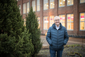 Tummansininen toppatakkinen mies seisoo ulkona. Taustalla näkyy punatiilinen rakennus ja sivulla jotain tuijaa tai muuta havukasvia.