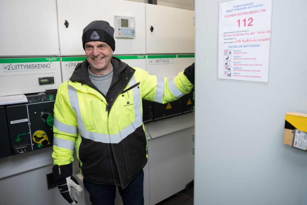 Huomiotakki päällä oleva mies nauraa iloisesti. Hän on jossain teknisessä tilassa, takana näkyy tekstejä, joissa lukee mm. liittymiskenno. Etualalla on ohjeet hätätilanteiden varalle.