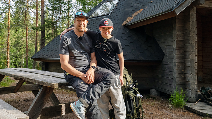 Mies ja poika ovat retkellä. Pojan käsi on isän ympärillä, takana näkyy kotaa.