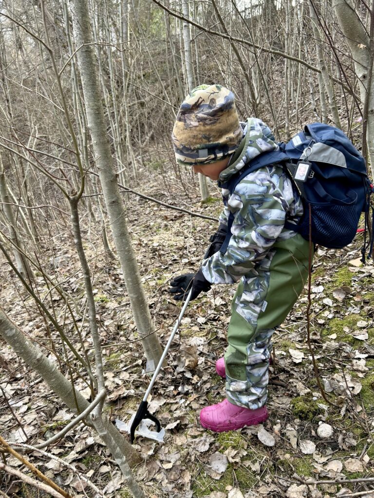 Pipopäinen lapsi kerää reppu selässä roskia luonnosta. On kevät, sillä puissa ei ole vielä lehtiä.