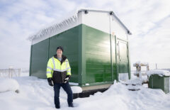 Mies hymyilee vihreä-valkoisen rakennuksen edessä. Rakennus on melko pieni ja uusi, sekä sen katolla ja ympärillä on paljon lunta.