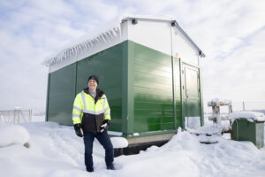 Mies hymyilee vihreä-valkoisen rakennuksen edessä. Rakennus on melko pieni ja uusi, sekä sen katolla ja ympärillä on paljon lunta.