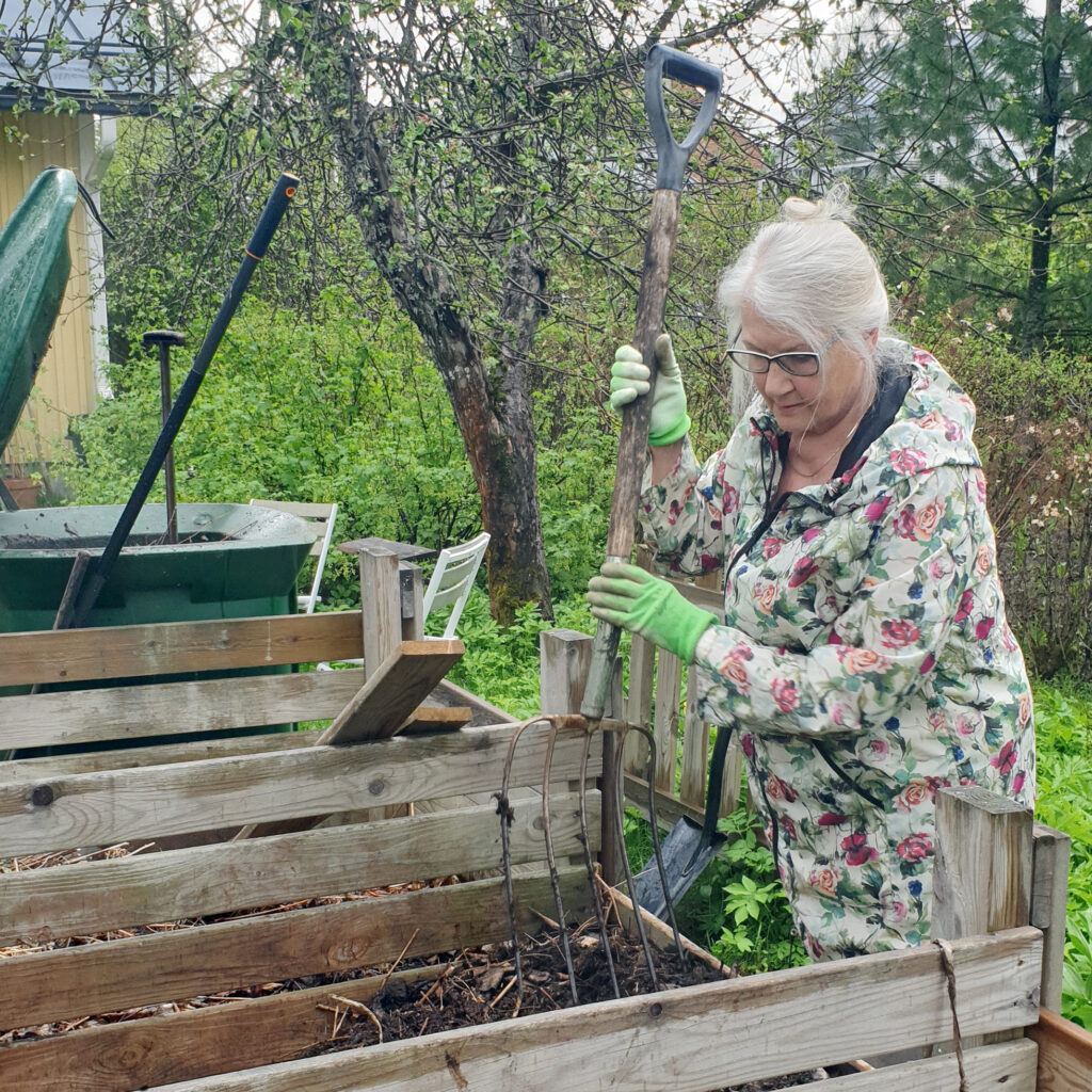 Harmaatukkainen nainen möyhii kompostoria. Hänellä on silmälasit ja päällään kaunis kukkatakki.