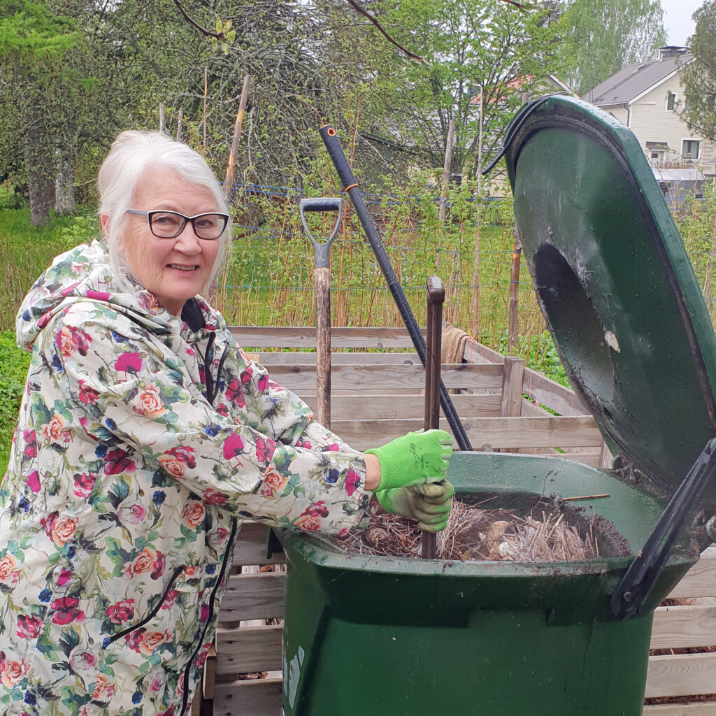 Harmaatukkainen nainen möyhii kompostoria. Hänellä on silmälasit ja päällään kaunis kukkatakki.