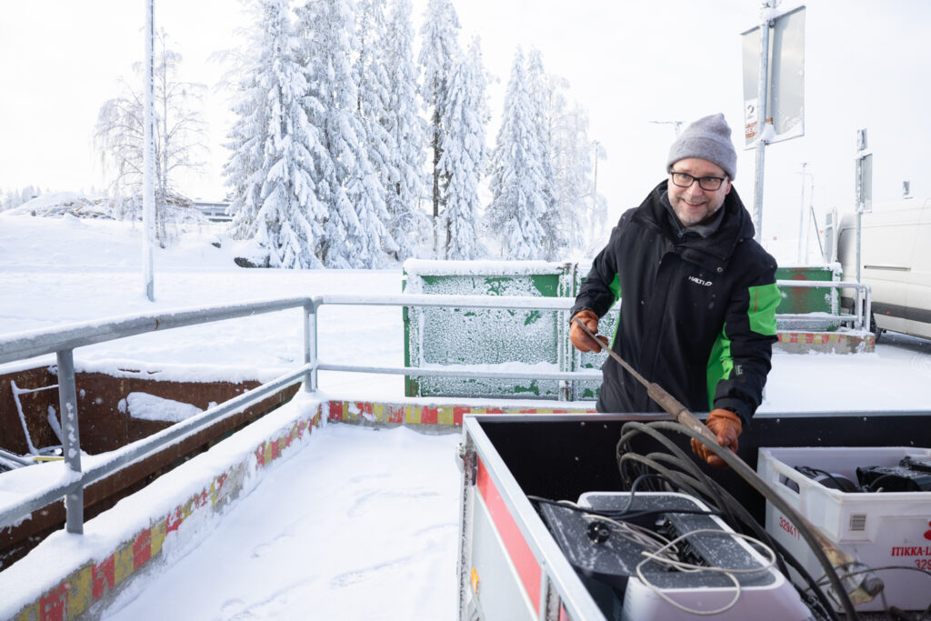 Pipopäinen mies ottaa peräkärryn lavalta jotain metalliromua. Takana näkyy talvinen maisema ja metallikaidetta sekä vihreitä lajittelulavoja.