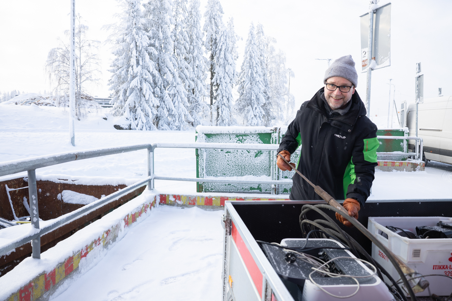 Pipopäinen mies ottaa peräkärryn lavalta jotain metalliromua. Takana näkyy talvinen maisema ja metallikaidetta sekä vihreitä lajittelulavoja.