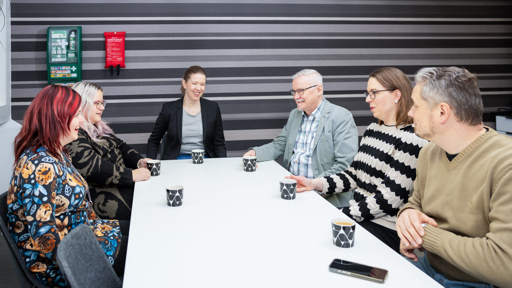 Ihmisiä kahvitauolla jossain toimistotilassa. Takana näkyy raitaseinäää ja ihmisiä naurattaa.
