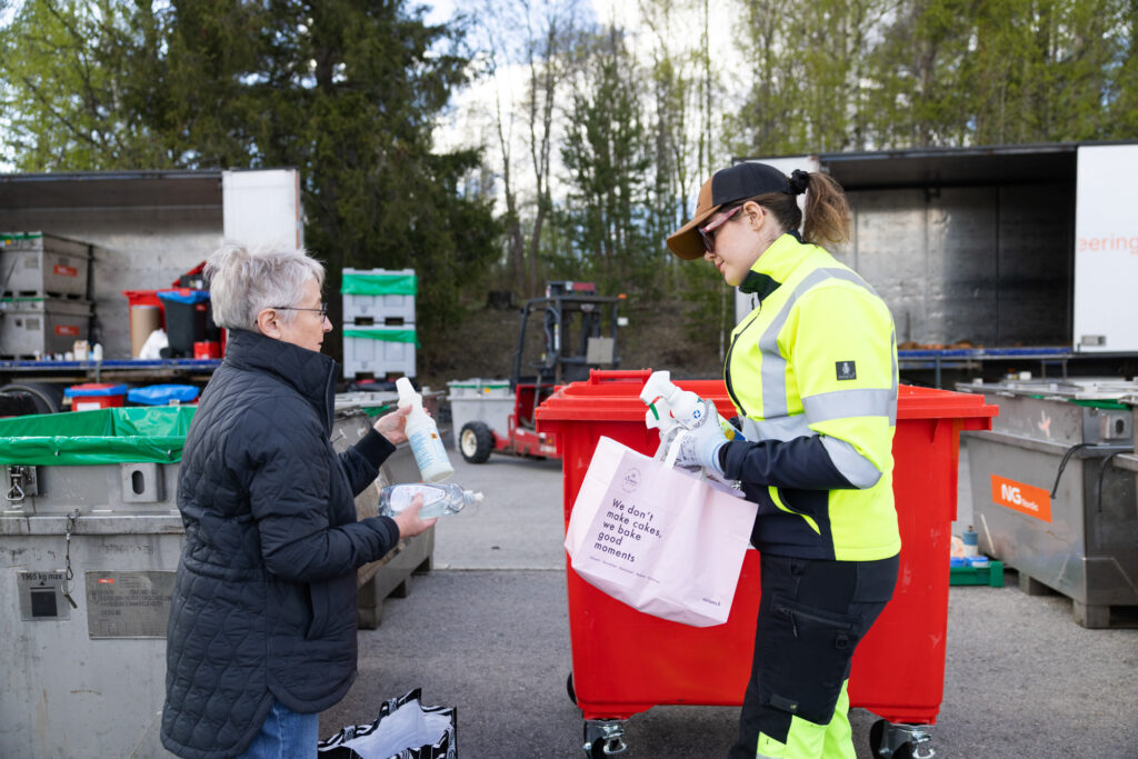 Mustatakkinen nainen ojentaa jonkinnäköistä purnukkaa nuorelle naiselle, jolla on päällään keltaiset huomiovaatteet. Takana näkyy jonkinlaisia astioita ja ajoneuvoa.