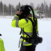 Joku huomiovaatteissa oleva henkilö laittaa päähänsä jonkinlaista maskia tai koneistoa.