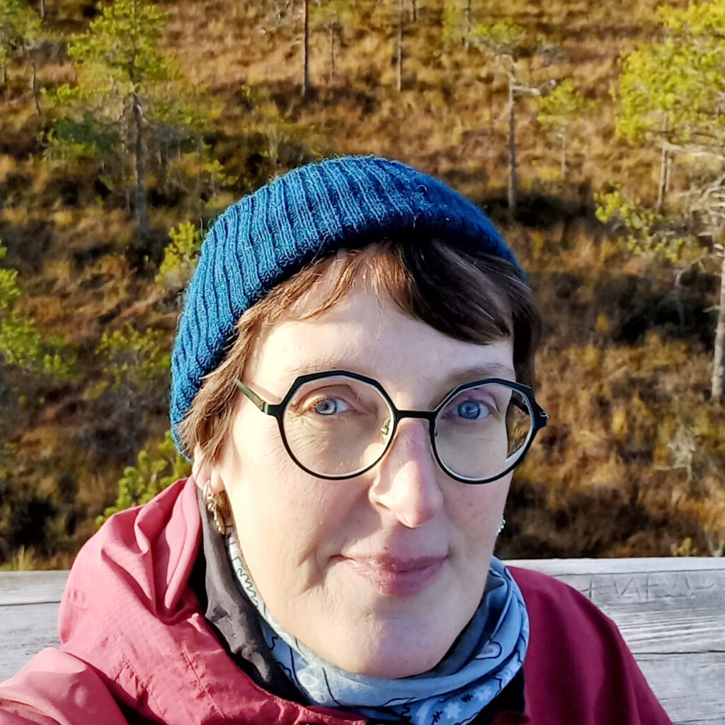 Silmälasipäinen nainen katsoo kameraan ja hymyilee vienosti. Hänellä on päässään sininen pipo ja takana näkyy suomaista luontoa.