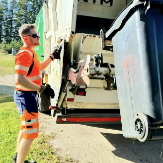 Huomiovaatteissa oleva mies seisoo jäteauton vieressä ja tyhjentää jäteastian sisältöä autoon.