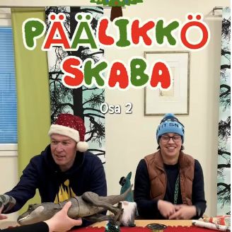 Tonttulakkipäinen mies ja sinipipoinen nainen istuvat vierekkäin. Heidän päälllään lukee teksti "Päällikköskaba, osa 2".