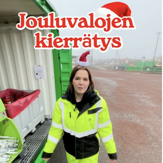 Nainen seisoo vihreä-valkoisen jätekontin luona ja hänen yläpuolellaan lukee "jouluvalojen kierrätys".