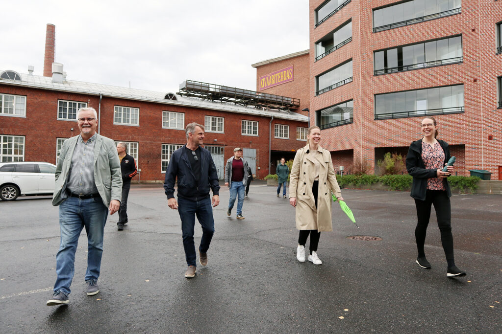 Seitsemän henkilöä kävelee punatiilisten rakennusten keskellä. Heitä hieman naurattaa.