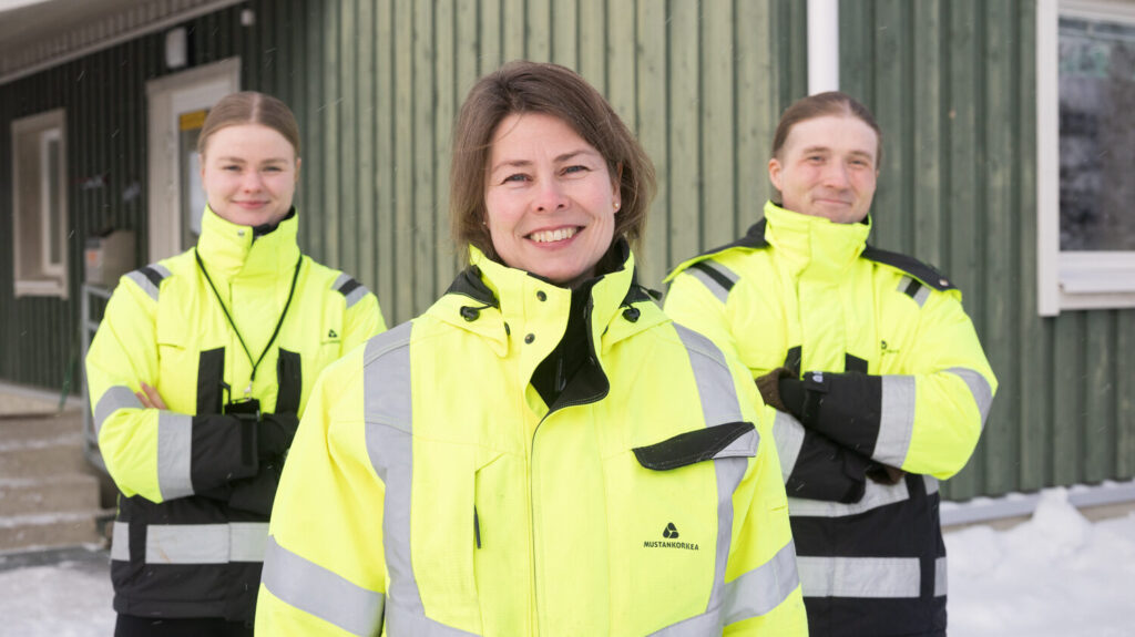 Ryhmäkuvassa on etualalla nainen ja hänen takanaan hieman nuoremmat nainen ja mies. Kaikilla on päällään keltaiset huomiovaatteet ja takana näkyy vihreää rakennusta.