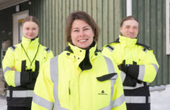 Ryhmäkuvassa on etualalla nainen ja hänen takanaan hieman nuoremmat nainen ja mies. Kaikilla on päällään keltaiset huomiovaatteet ja takana näkyy vihreää rakennusta.
