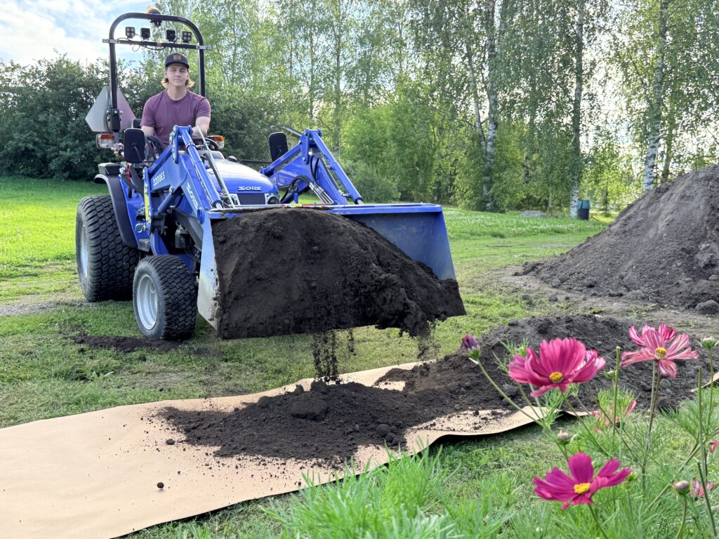 Mies kaataa pienellä kauhakuormaajalla multaa kohopenkkiin. Taustalla näkyvät puut ja omakotitalon piha.
