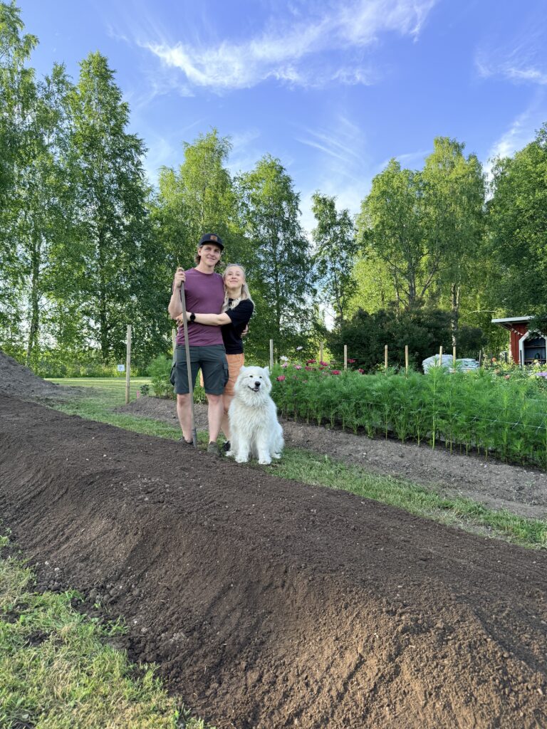Pariskunta sekä valkoinen koira seisovat puutarhassa kohopenkin takana ja katsovat kameraan. Taustalla näkyy puita ja keli on poutainen.