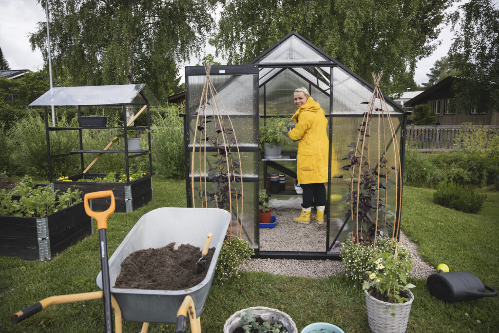 Kasvihuoneessa on keltatakkinen nainen. Etualalla näkyvät myös kottikärryt. Naisella on päällään keltainen sadetakki.