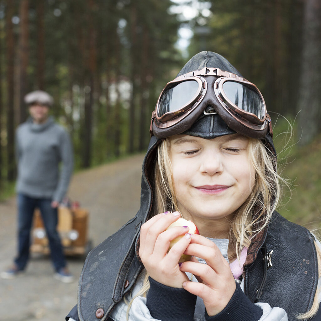 Lapsella on päällään hieman hassut varusteet ja hän syö omenaa. Taustalla näkyy isä vetävän jotain perässään.