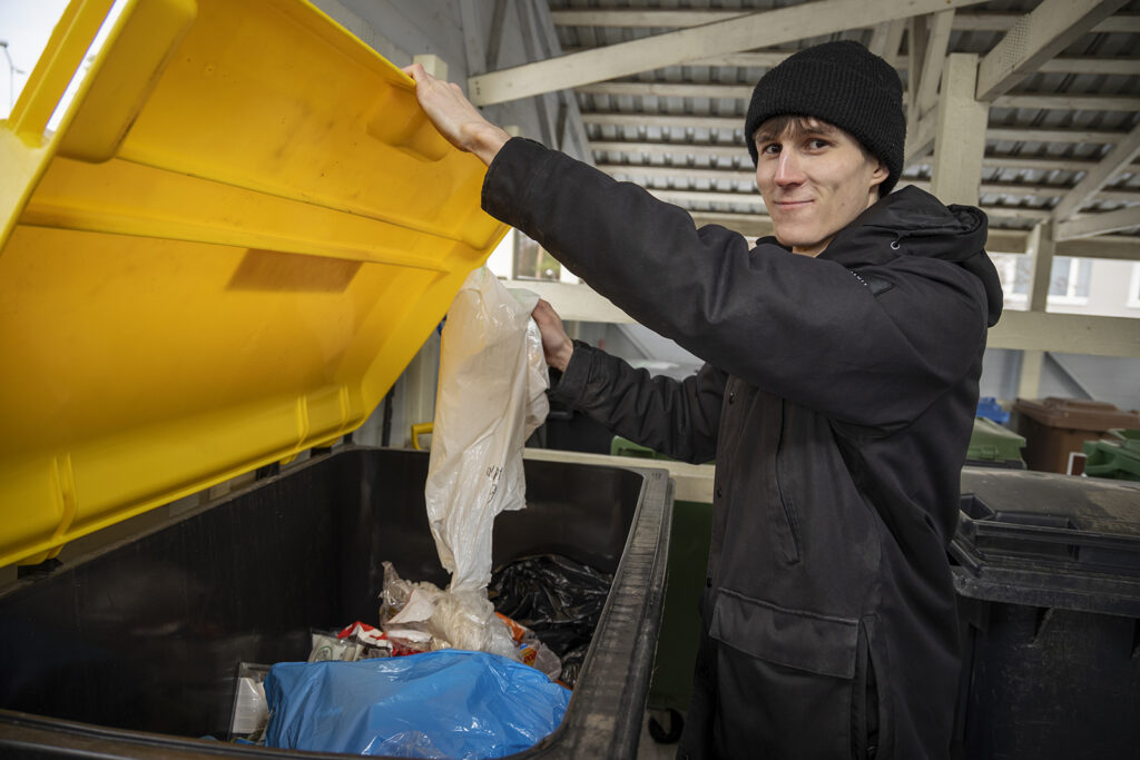 Nuori mies kaataa keltaiseen jäteastiaan muoviroskia. Hänellä on tummat vaatteet ja musta pipo.